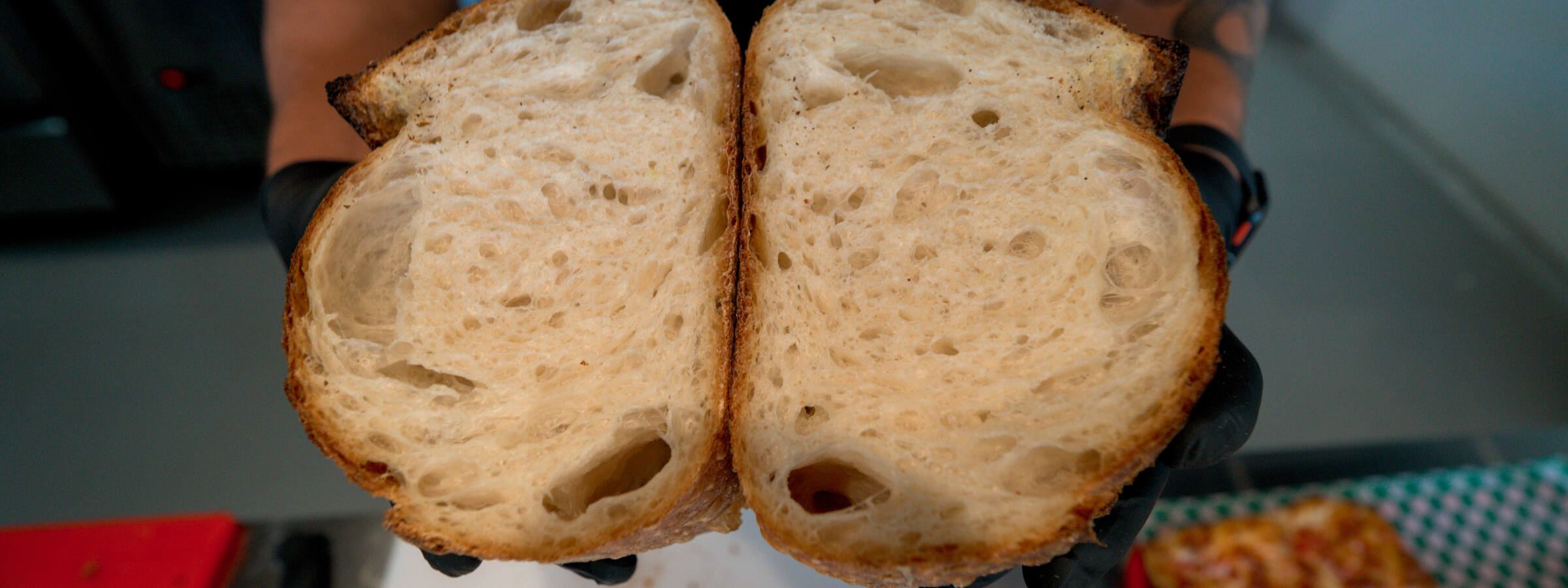 Split sourdough loaf held open in black-gloved hands, showing its airy crumb and golden-brown crust